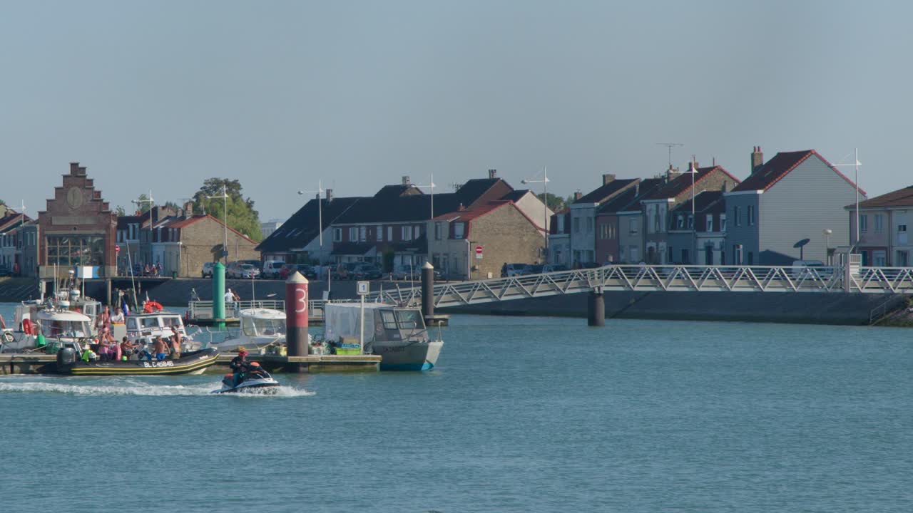 Jet ski crosses calm harbor near marina and waterfront village in bright daylight, static shot