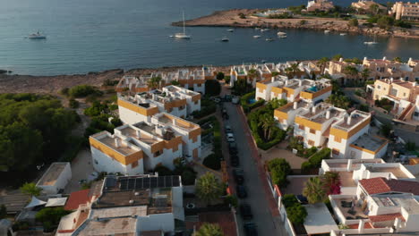 Aerial-view-of-rooftop-of-beautiful-houses-near-the-beach-and-shore-with-boats-and-yachts-parked-surrounded-by-ocean-and-mountain-in-Ibiza-in-Spain-during-an-early-morning