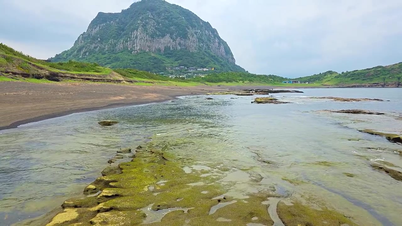 Vista dal basso di rocce di tufo coperte di muschio sulla spiaggia di Yongmeori con il monte vulcanico Sanbangsan che si erge sopra la costa sabbiosa sull'isola di Jeju