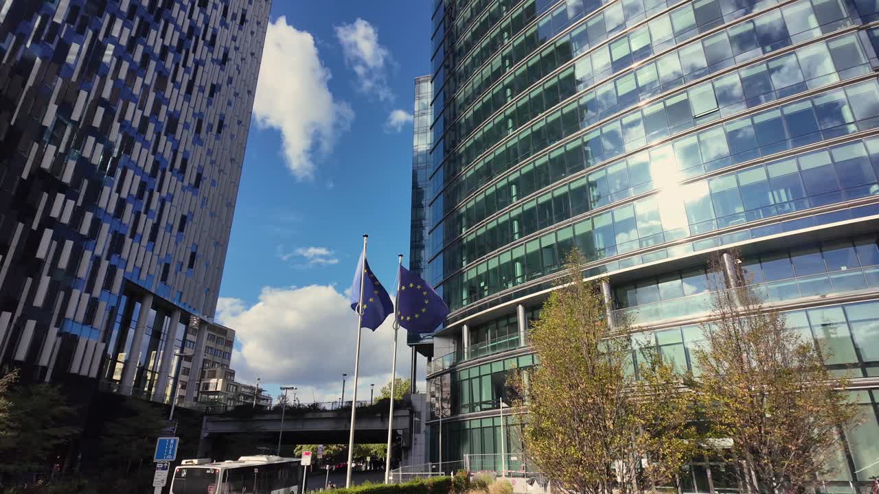 European Union flags flutter under a blue sky between the Lex Building and modern glass tower on Rue de la Loi in Brussels’ EU district