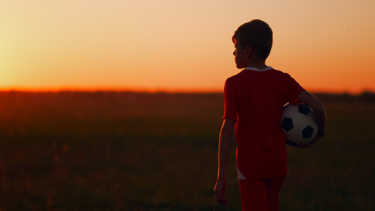 el niño está en el campo con la pelota en las manos mirando la puesta de sol y soñando con una carrera futbolística. la cámara sigue al niño