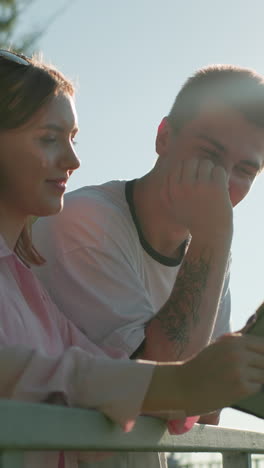 chica de camiseta rosa sonriendo mientras le muestra a su hermano algo en su tableta, él sacude la cabeza con una mano y se cubre la boca con la otra mientras el sol brilla sobre ellos