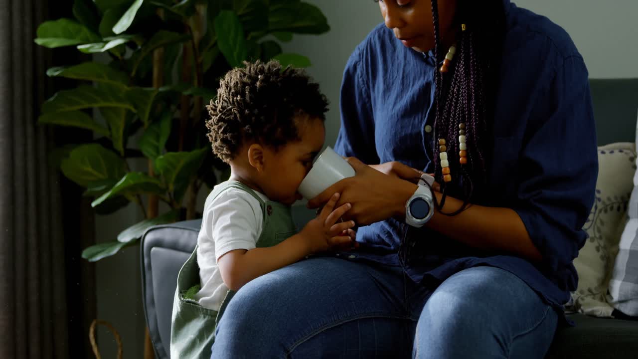 vista frontal de una joven madre negra alimentando leche de vidrio a su hijo en un hogar cómodo 4k