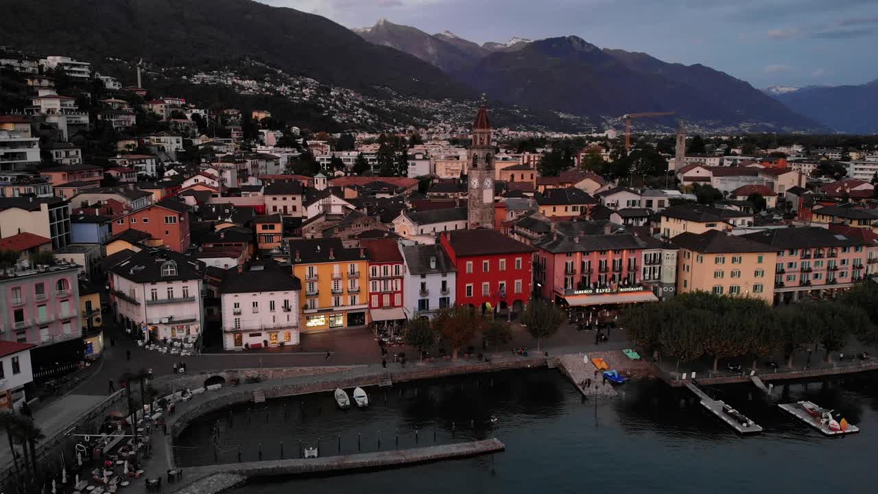 vista aérea de la orilla del lago maggiore en ascona, suiza con un descenso que revela barcos y los alpes detrás de la torre de la iglesia y una casa colorida a lo largo del paseo junto al lago