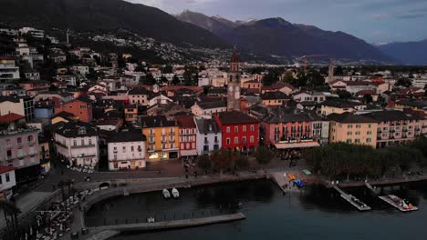 aerial view of the shore of lago maggiore in ascona, switzerland with a descent revealing boats and the alps behind the church tower and colorful house along the lakeside promenade