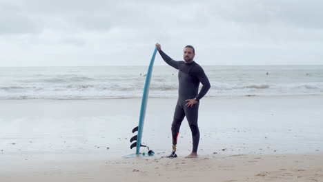 long shot of a handsome man in wetsuit with bionic leg standing on seashore with surfboard, smiling at the camera and showing shaka sign