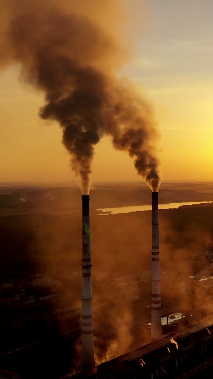 Metallurgy plant in nature at orange sunset. Dark smoke of industrial factory pollute the atmosphere. Evening background of a factory near the river. Vertical video
