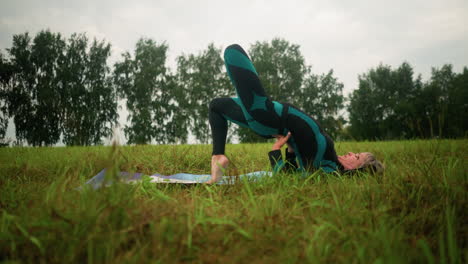 middle-aged woman lying on a yoga mat practicing bridge pose with hands supporting her waist in a vast grassy field under cloudy skies, with trees lined in the distance