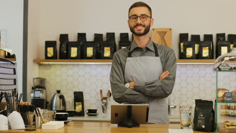 barista caucásico con barba y anteojos usando la tableta, luego mira la cámara y sonríe con los brazos cruzados en la cafetería