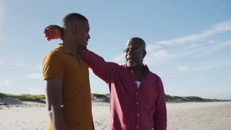 padre afroamericano de pie en la playa poniendo la mano en el hombro de su hijo adolescente y hablando