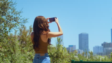 unknown brunette take picture cityscape standing on green park back view.