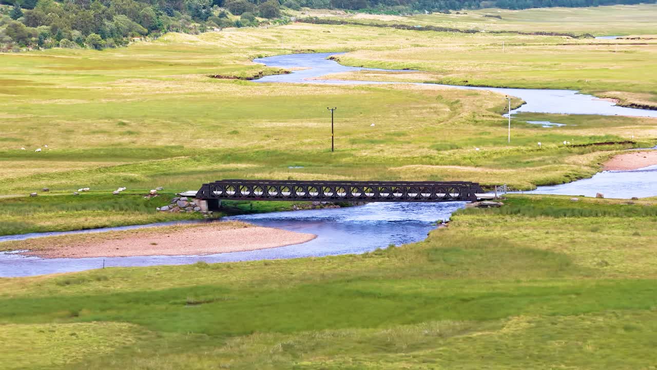 Drone pans across tranquil river, metal bridge, and green valley under soft daylight in Scotland