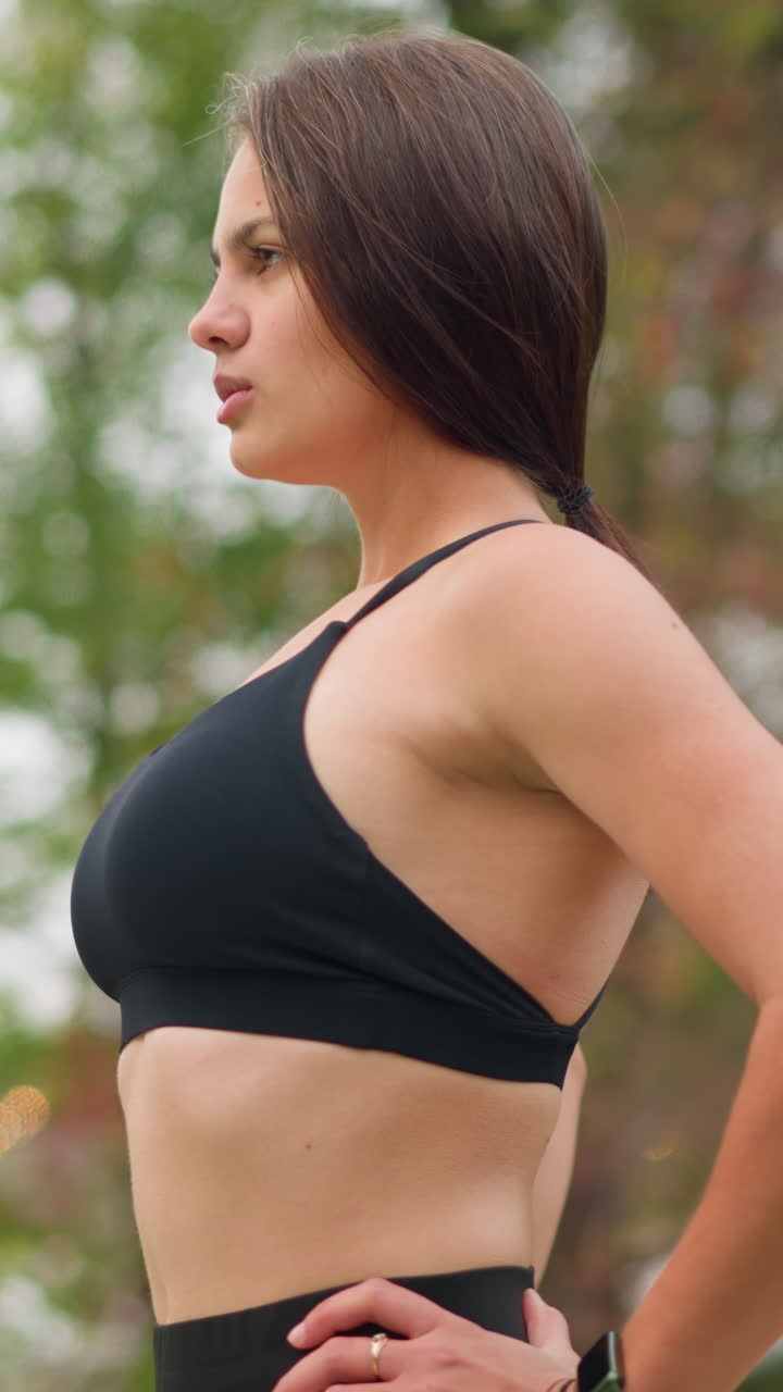 Girl outdoor in black singlet standing and breathing in deeply with hand on waist, calm and focused expression, healthy posture, feeling refreshed, natural beauty, fitness moment, outdoor breath