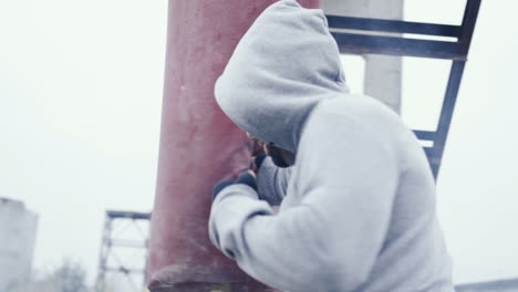 close-up view of caucasian man in grey hoodie hitting a punching bag outdoors an abandoned factory on a cloudy morning