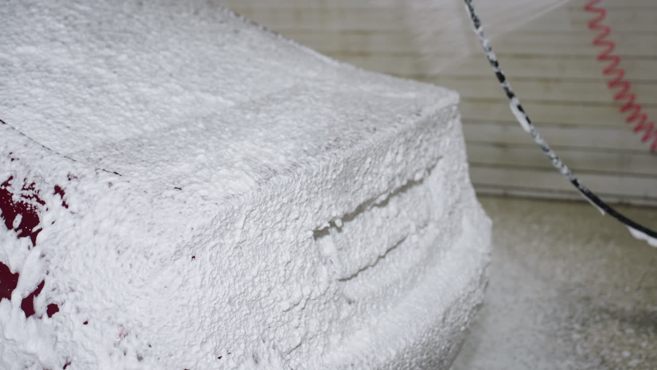 Side close up of professional car washing process showing dense white foam covering red vehicle surface as high pressure water spray from hose nozzle creates cleansing bubbles inside indoor garage