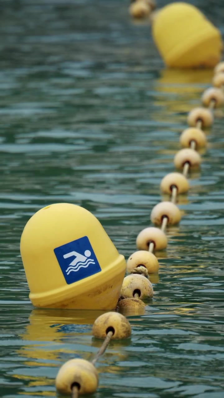 Yellow swimming buoy with a blue sign marking a safe area, floating on green water, part of a rope barrier