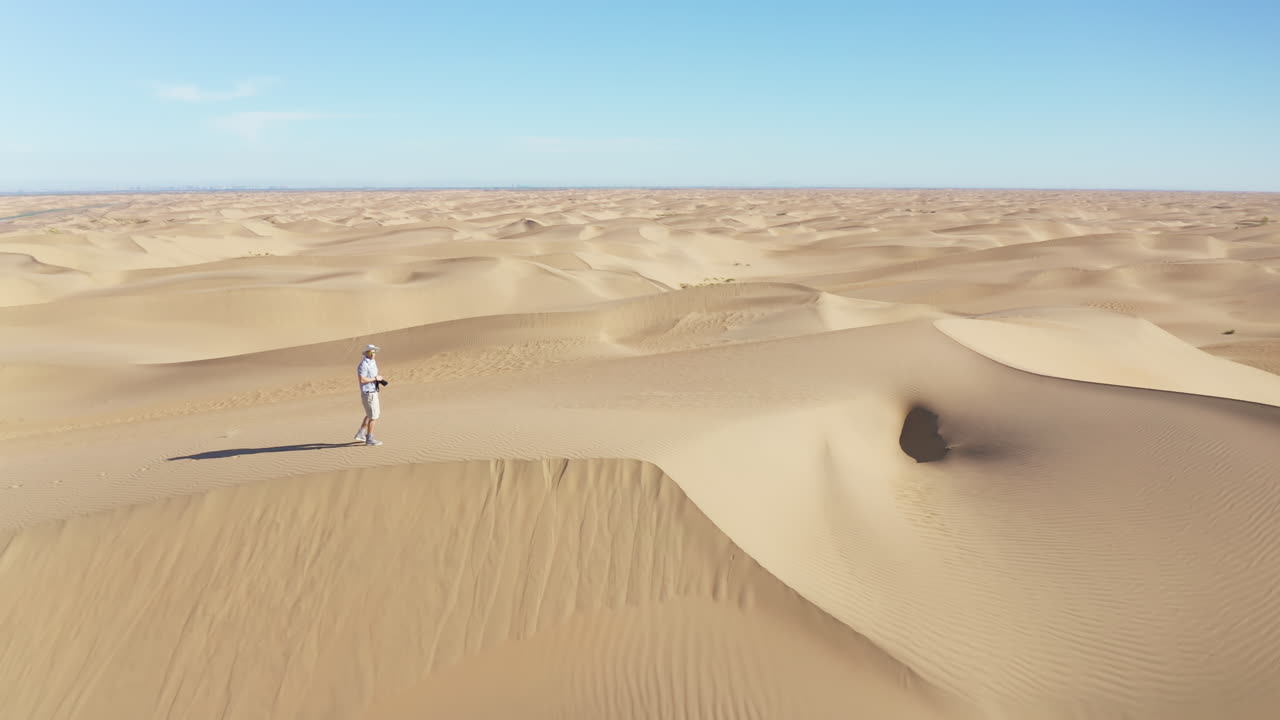 imágenes cinematográficas de un hombre caminando por una duna de arena empinada en el desierto, sosteniendo una cámara y tomando fotos