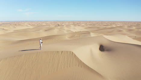 cinematic drone footage of a man walking up a steep sand dune in the desert, holding a camera and taking photos