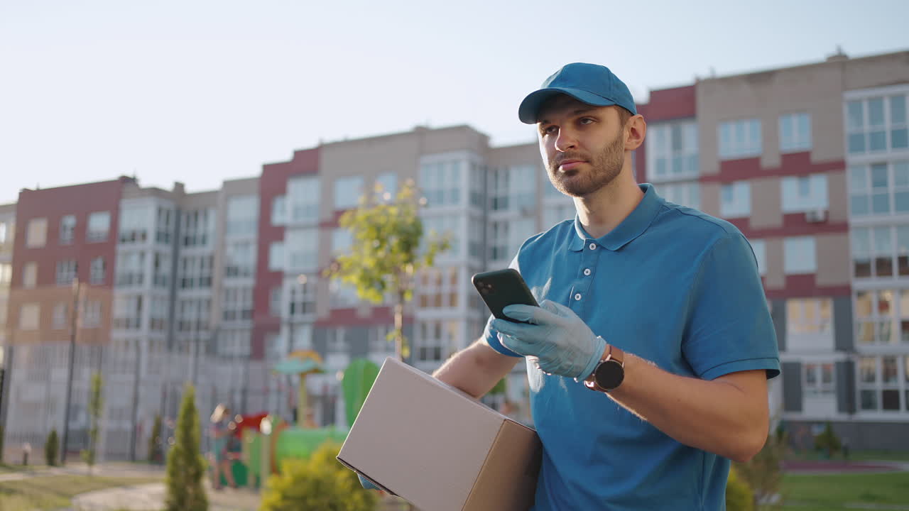joven caucásico trabajador de entrega de hombre guapo con gorra azul caminando por la calle y llevando una caja de cartón mientras usa un teléfono inteligente buscando una ruta. mensajero masculino con paquetes tocando y enviando mensajes de texto por teléfono.