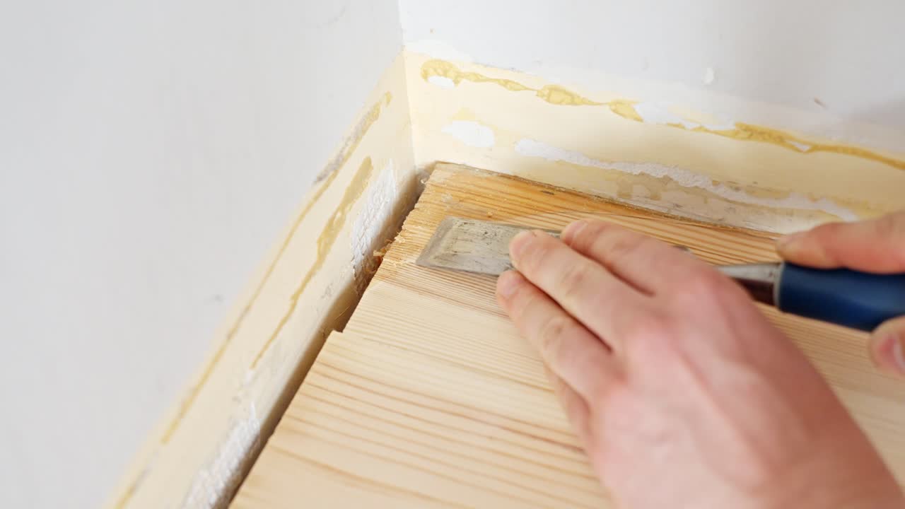 Close-up of hands using a flat scraper to remove old varnish or paint from a pinewood floor edge, preparing the surface for refinishing, static view, real time