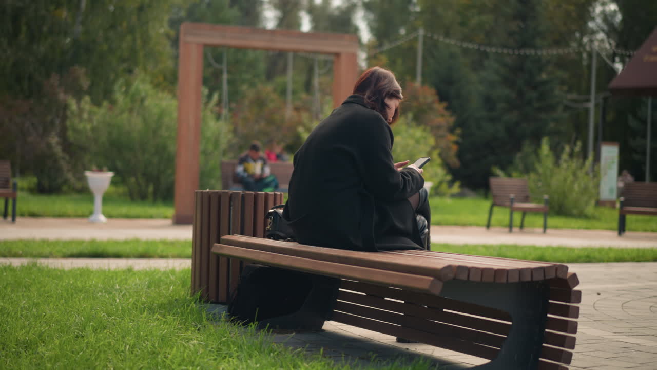 공원 벤치에 앉아 스마트폰에 집중하는 여성, 검은 코트와 부츠를 입고, 울창한 녹색 나무로 둘러싸여, 도시 공원의 평화로운 야외 분위기, 흐릿한 스 의자 전망