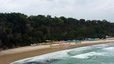 Dolly-out-aerial-drone-shot-of-the-famous-tropical-Madeiro-beach-near-Pipa,-Brazil-in-Rio-Grande-do-Norte-with-calm-waves-for-surfing,-colorful-umbrellas,-and-surrounded-by-large-exotic-cliffs