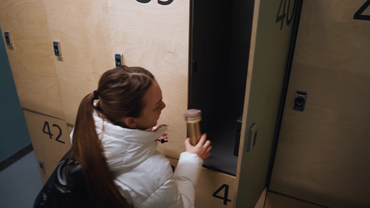 White woman opening numbered locker retrieving bag and securing coat puffer jacket, ponytail, plywood lockers, quick glance, purposeful stride through teal corridor of urban sports facility commuter