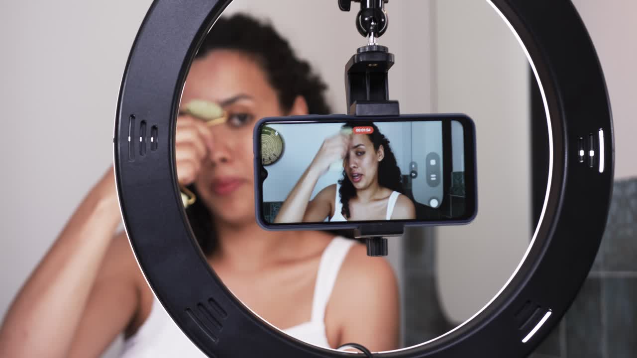 African American woman in bathroom using roller while phone recording with ring light, giving tips