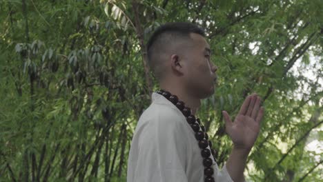 a man with short hair and large wooden beads around his neck meditates outdoors in a bamboo forest, focusing with one hand raised