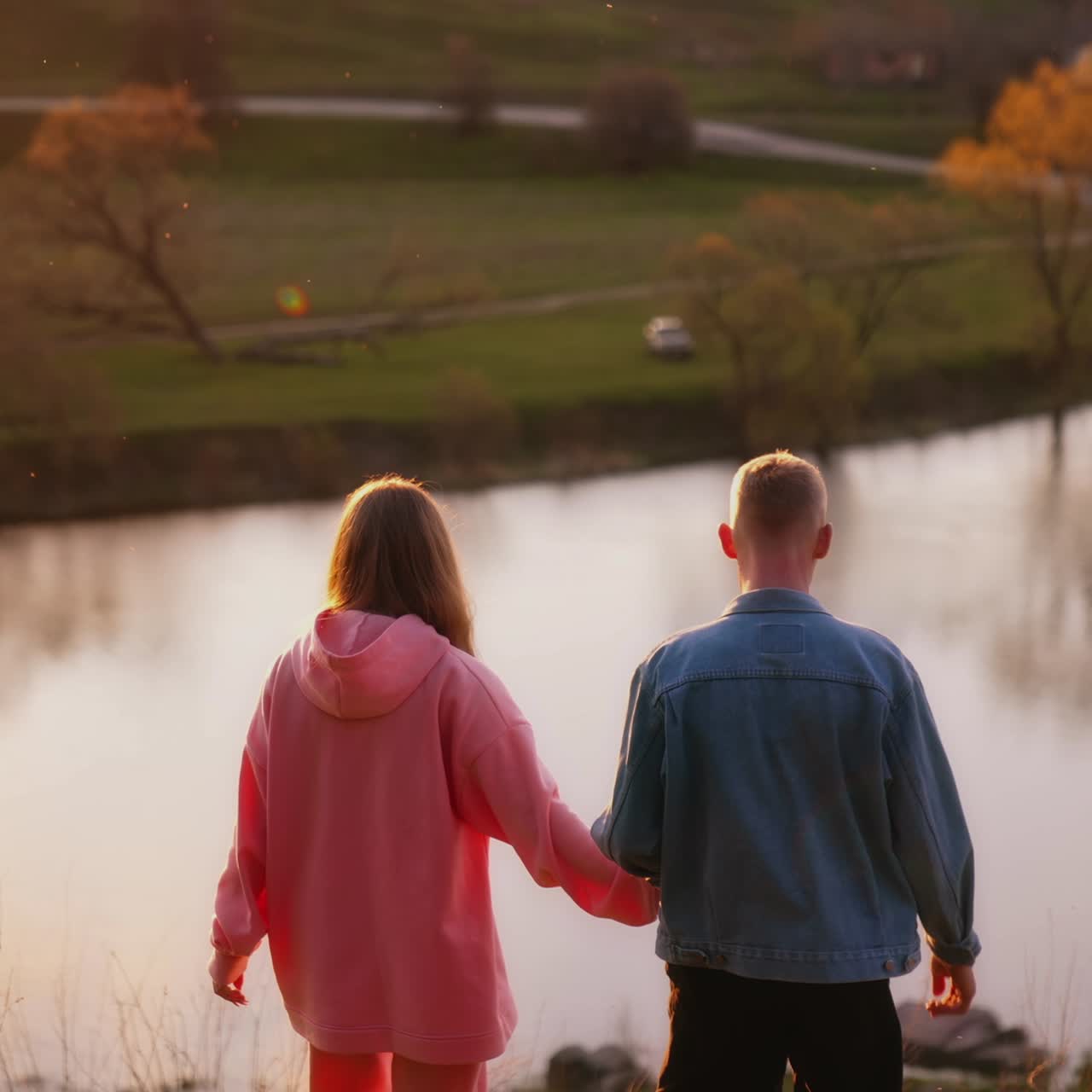 Happy young people walking in nature at sunset. Beautiful girl and blond guy holding hands and walk near the river in the evening