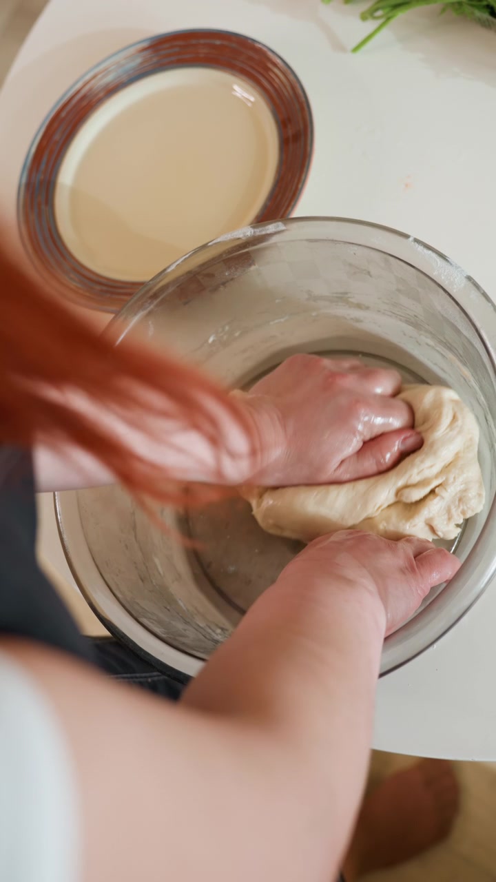 Mãos de cima amassando massa elástica dentro de uma tigela de vidro transparente na bancada da cozinha. Pressionar, dobrar e esticar rápidos e rítmicos criam uma cena animada de panificação caseira com luz natural suave.
