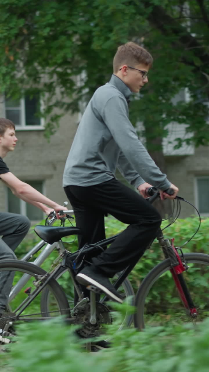 두 친구가 울창한 초록색 지역을 가로질러 자전거를 타고 있는 측면 전망, 한 명이 회색 에 서 있고, 다른 한 명이 검은 에 앉아 있으며, 배경에는 초록색, 흐릿한 건물이 있습니다.