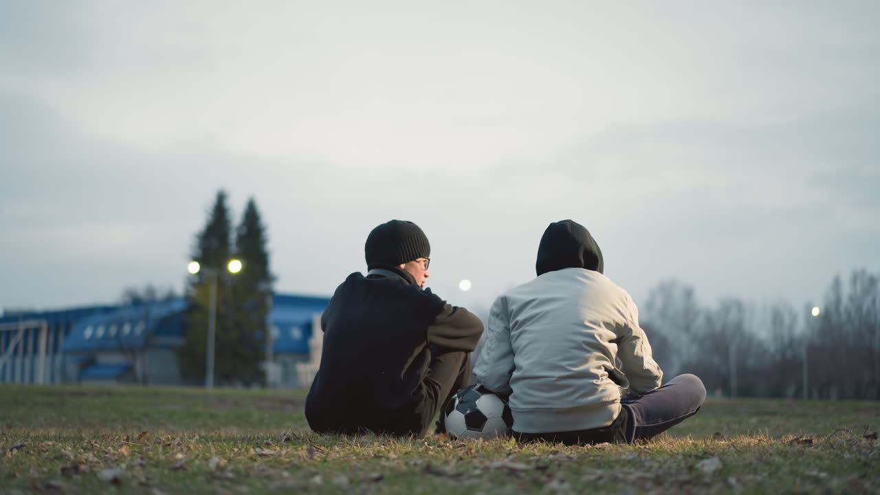 Two boys are seated on a grassy field, one wearing a gray jacket resting his left hand on a soccer ball, with blur light view and building in the background