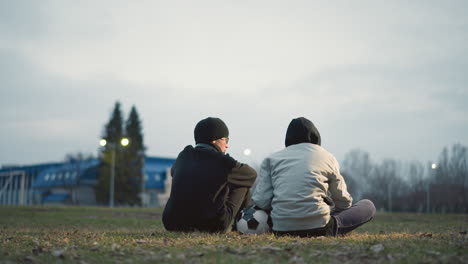 two boys are seated on a grassy field, one wearing a gray jacket resting his left hand on a soccer ball, with blur light view and building in the background