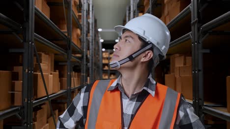 close up of asian male engineer with safety helmet in the warehouse with shelves full of delivery goods. standing with arms akimbo looking around, checking the stock on racks