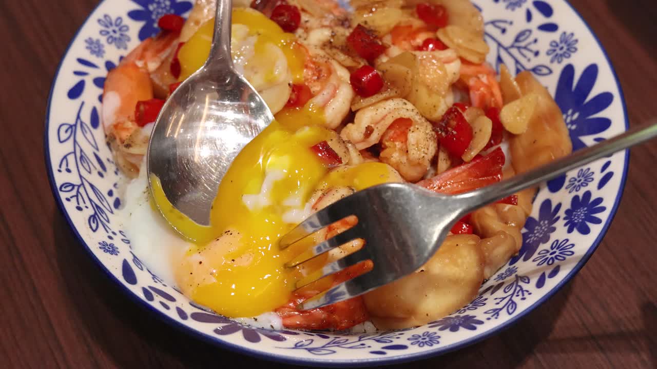 A spoon and fork break a soft-boiled egg, releasing yolk over garlic shrimp rice with chili in a patterned bowl under warm indoor lighting