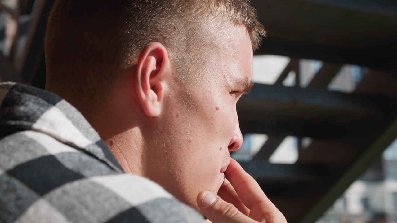 Close up of young man exhaling smoke while smoking cigarette under staircase with urban structure in soft focus background and sunlight illuminating face and side profile in contemplative expression