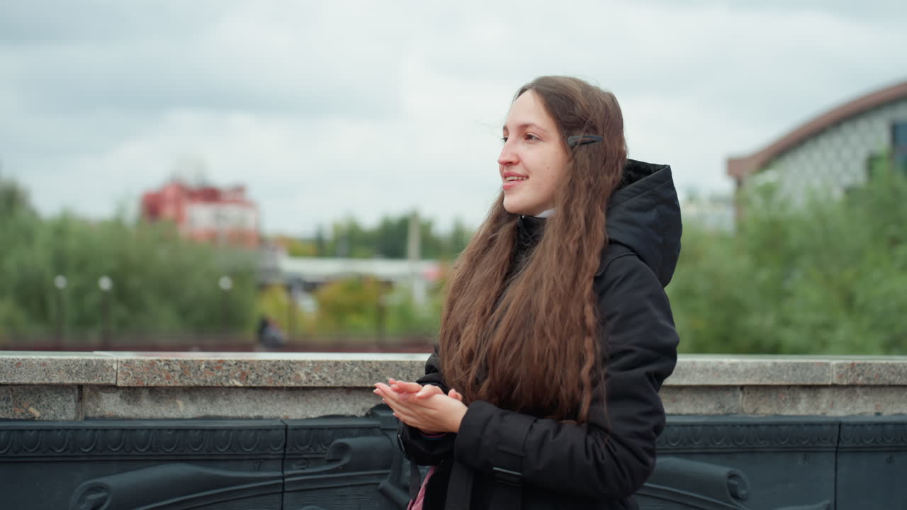 Donna caucasica sorridente su un ponte sul fiume durante una passeggiata contemplativa, con cappotto e cappuccio neri, lunghi capelli castani appoggiata alla ringhiera, cielo autunnale nuvoloso, architettura e vegetazione in lontananza, serena e spontanea