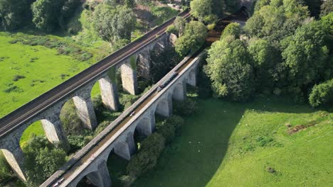 Barco-Estrecho-Y-Canoas-Cruzando-El-Canal-Sobre-El-Acueducto-De-Chirk,-Viaducto-Ferroviario-En-El-Fondo---Drone-Aéreo-Gira-En-Sentido-Antihorario,-Retrocede-Y-Luego-Avanza---Frontera-Galesa-E-Inglesa,-23-De-Septiembre