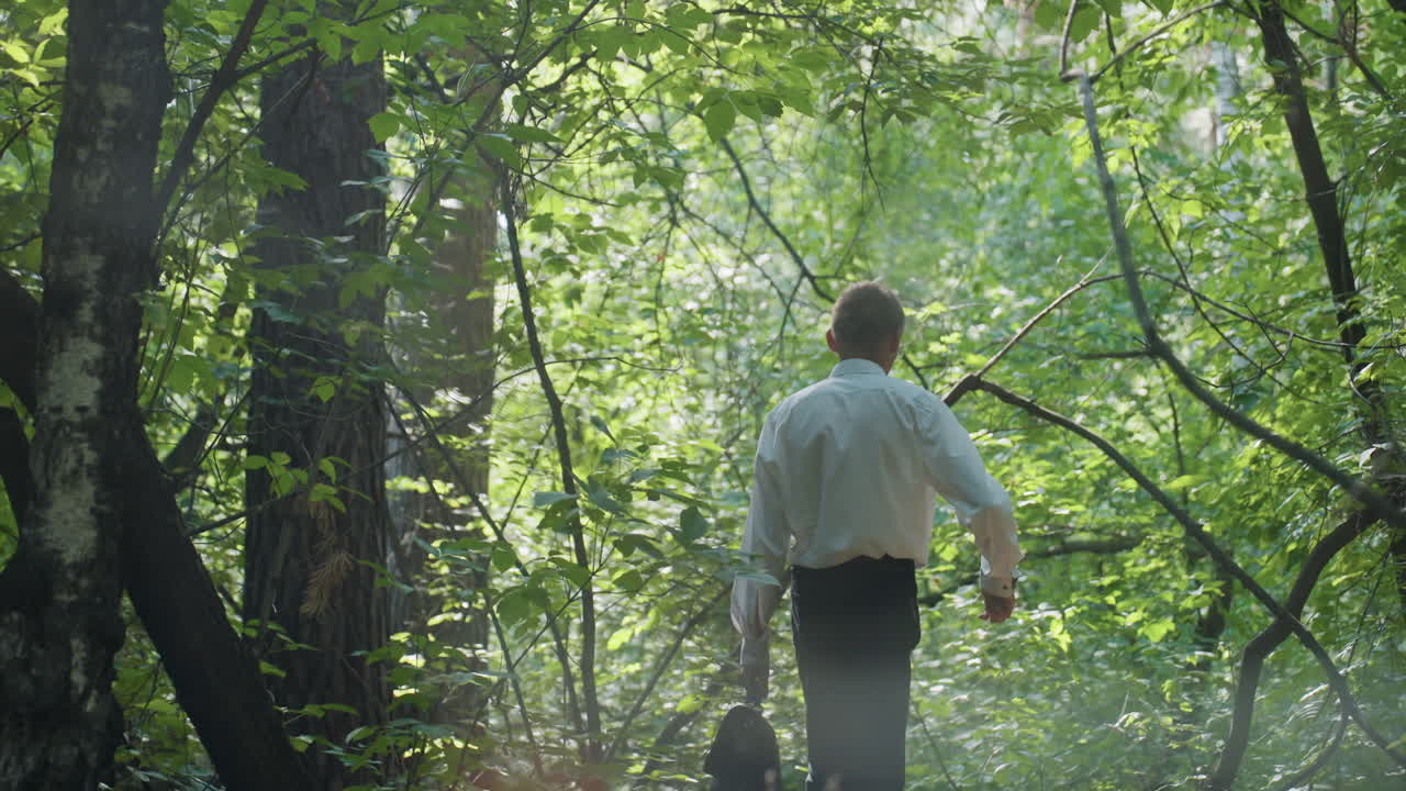 Biologist in white shirt and black trouser carrying backpack while bending carefully and walking through dense green forest under daylight, moving cautiously between trees and branches