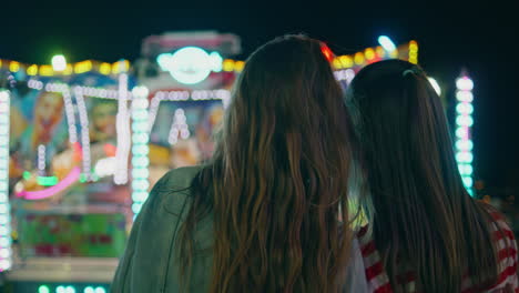 funfair girls watching illuminated carousel rear view. two happy teenage friends