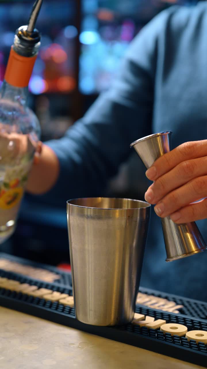 Barman che versa un distillato trasparente da una bottiglia in un jigger di metallo. Primo piano della misurazione di ingredienti liquidi usando un jigger sopra uno shaker sul bancone.