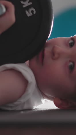 cute little boy with heavy barbell in hand turns head to camera at bright light. adorable toddler lies on floor in gym enjoying iintensive workout slow motion