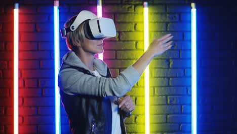 close up view of young blonde woman in vr glasses moving her hands in the air in a room with colorful neon lamps on the wall 1