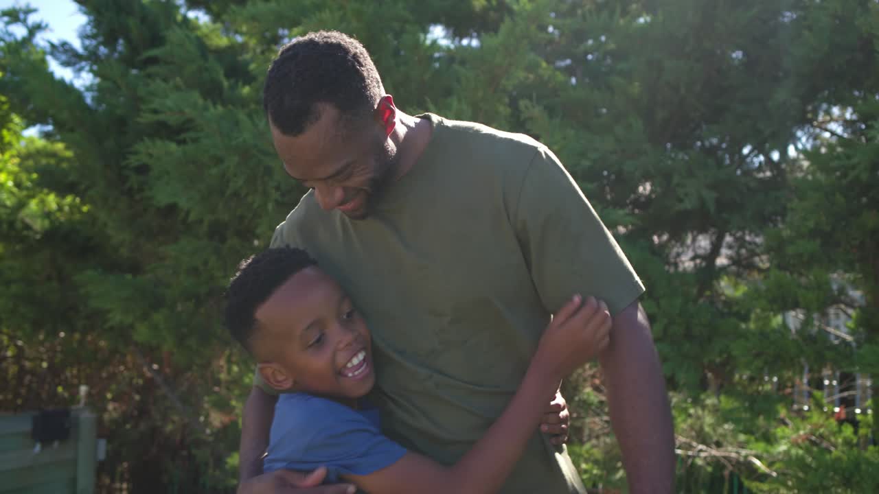 Afroamerikansk far og søn i baghaven; drengen taler, mens faren læner sig ned, roder i hans hår og knytter bånd.