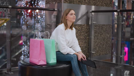 young woman seated in a mall holding a tablet while looking thoughtfully, surrounded by festive decor with glowing lights and shopping bags