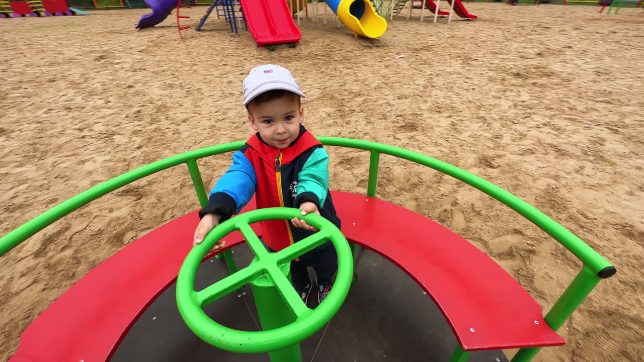 Lovely Caucasian toddler sitting on the merry-go-round. Happy kid holding by the green steering wheel. Close up.