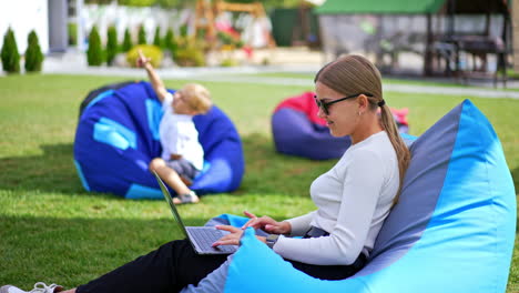 relaxed young lady wearing sunglasses working at laptop sitting in bean bag chair. little blond boy in chair at backdrop in blur.