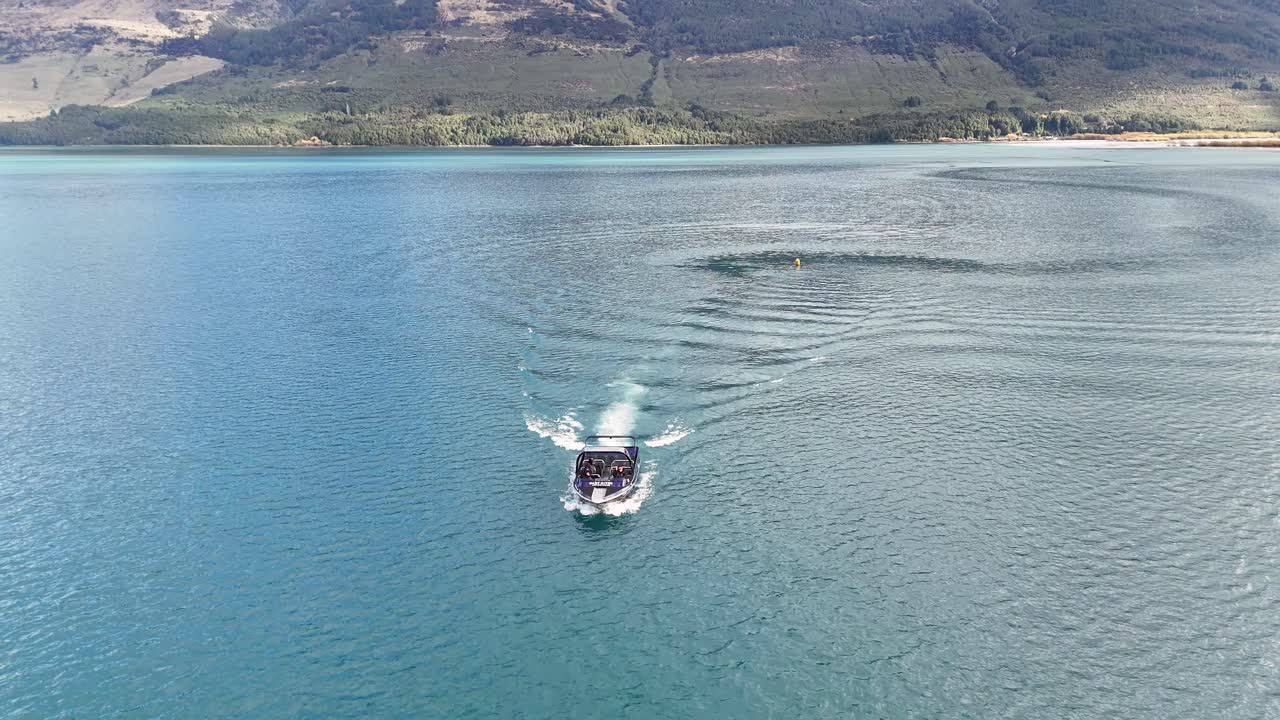 A speedboat navigates tranquil blue waters near Glenorchy, New Zealand, under clear skies with distant mountains in view
