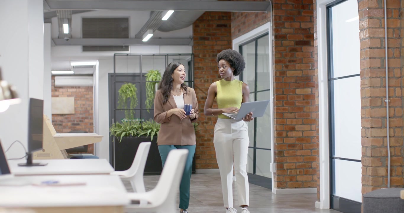 Diverse female coworkers starting discussion, walking along office aisle referencing laptop and mug